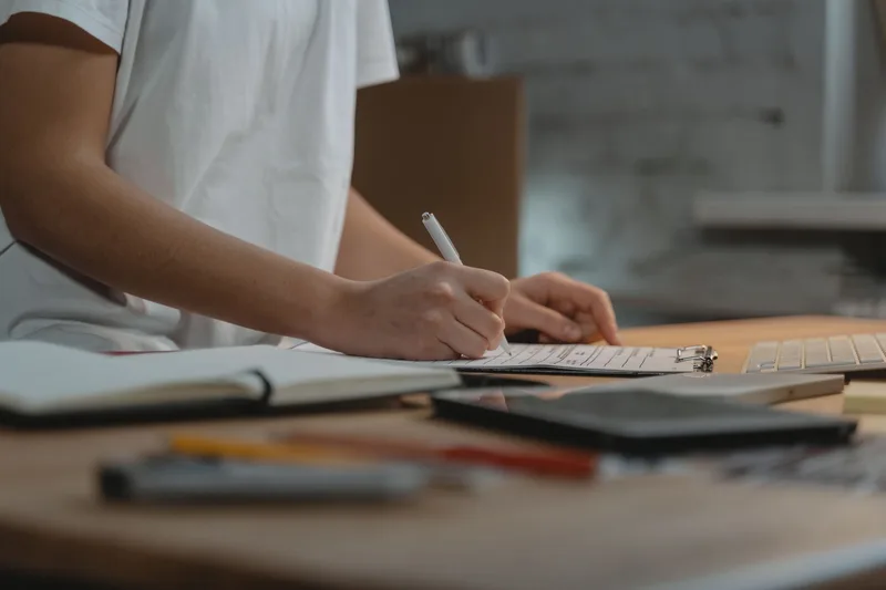 Person in a white T-shirt writing on a clipboard at a wooden desk, with a notebook, pens and a tablet beside it