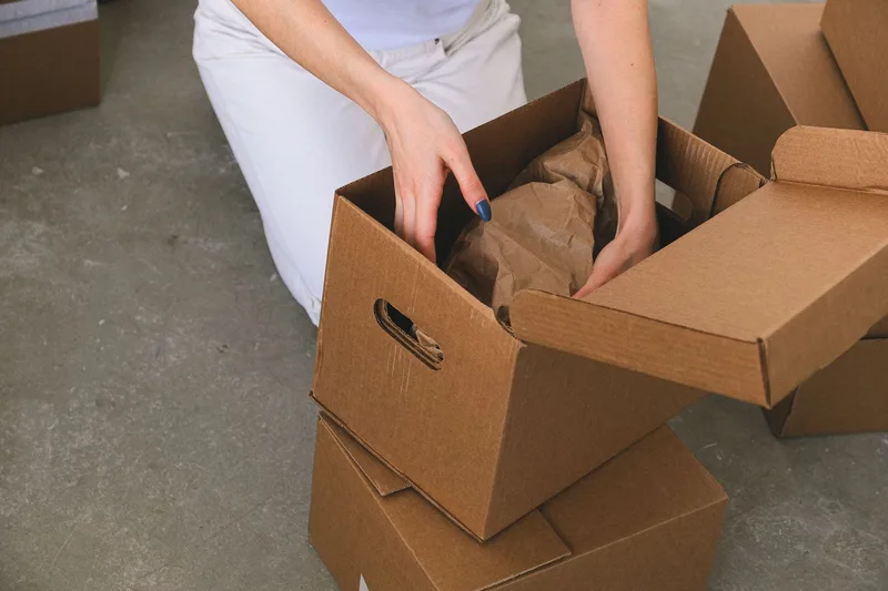 Person packing paper-wrapped items into a cardboard box, with more boxes stacked nearby