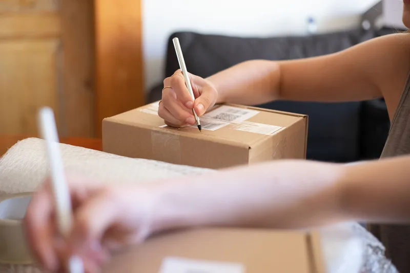 Hands writing on a cardboard parcel with a barcode label, with bubble wrap and tape beside it