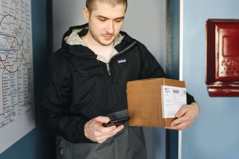Man holding a cardboard parcel with a shipping label while looking at his smartphone