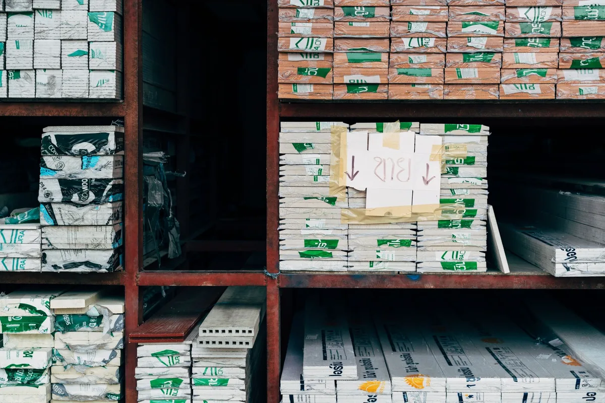 Steel shelves in a storeroom holding neatly stacked packs of building materials, with a handwritten label taped to one stack