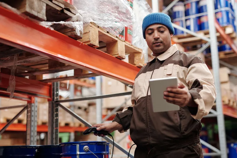 Warehouse worker in a blue beanie holding a tablet and a handheld barcode scanner beside pallet racking