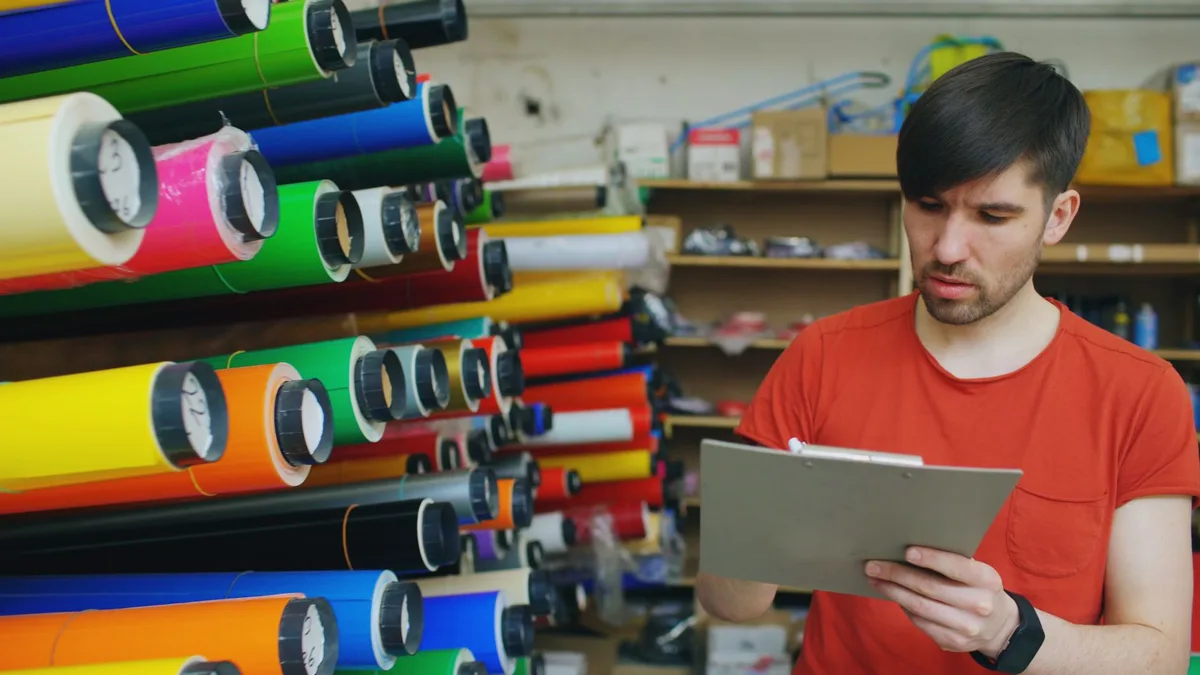 Man holding a clipboard to check stock beside racks of colourful material rolls in a storeroom