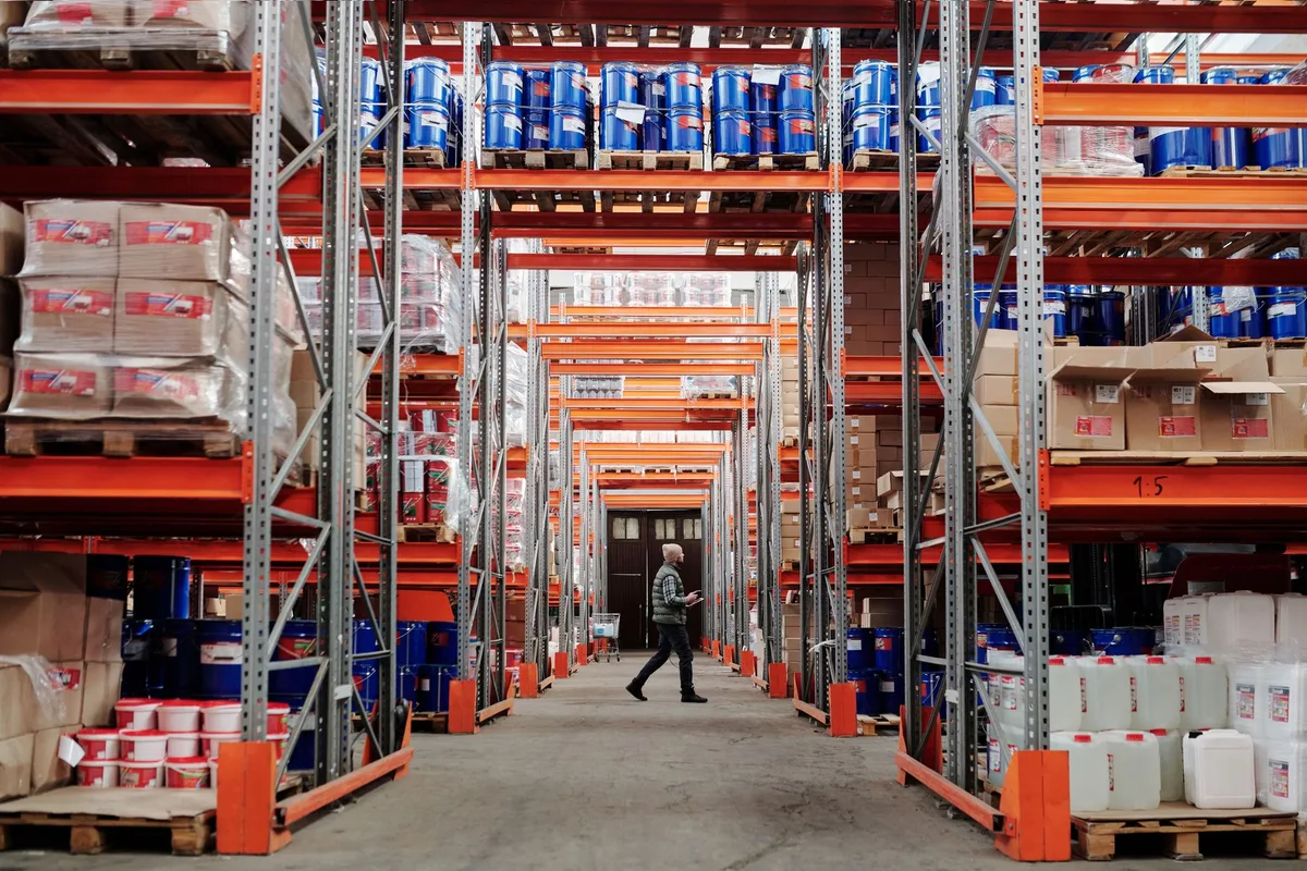 Warehouse aisle with orange racking holding boxes and containers, and one worker walking between the shelves