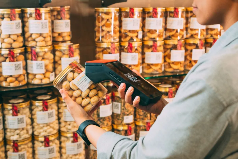 Shop worker scanning the QR label on a jar of cookies with a handheld scanner in front of shelves of labelled jars