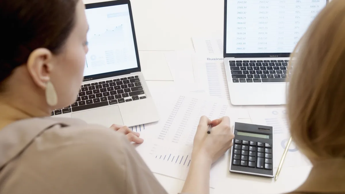 Two people at a desk reviewing printed figures next to two laptops showing a chart and a table of numbers, with a calculator between them
