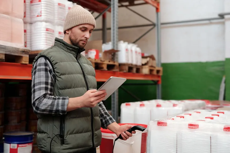 Warehouse worker in a vest holding a tablet and barcode scanner while checking stock near shelving stacked with product