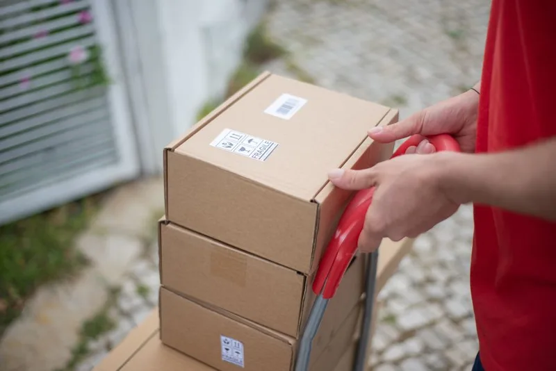 Hands holding a red hand truck loaded with stacked cardboard boxes carrying barcode and fragile labels