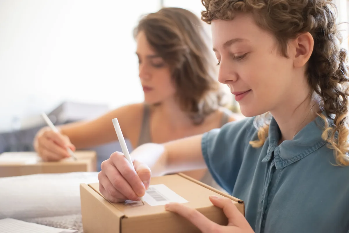 Two sellers hand-labeling parcel boxes with barcode stickers before shipping