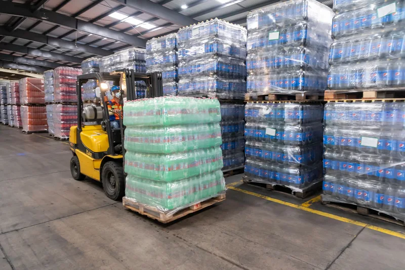 Forklift operator moving stacked pallets in a busy warehouse