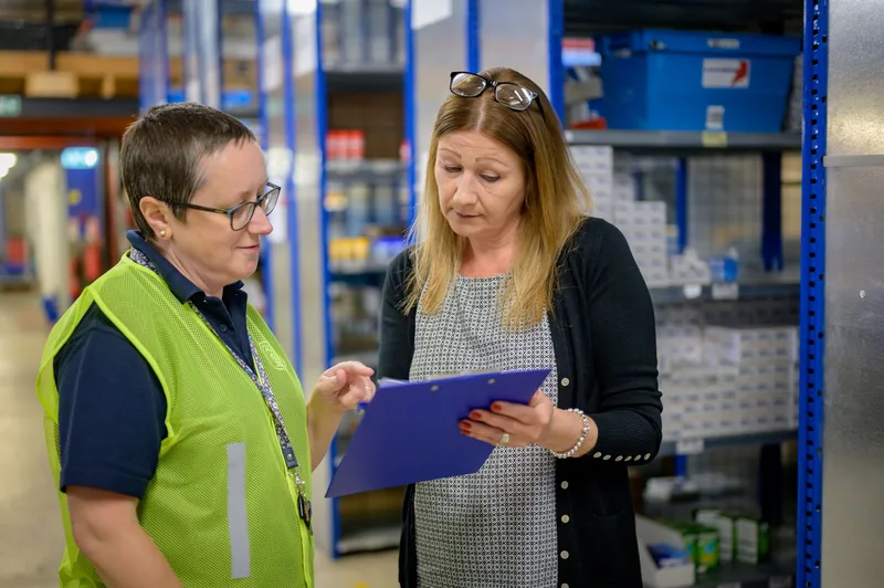 Two warehouse staff reviewing an inventory checklist on a clipboard near metal shelves