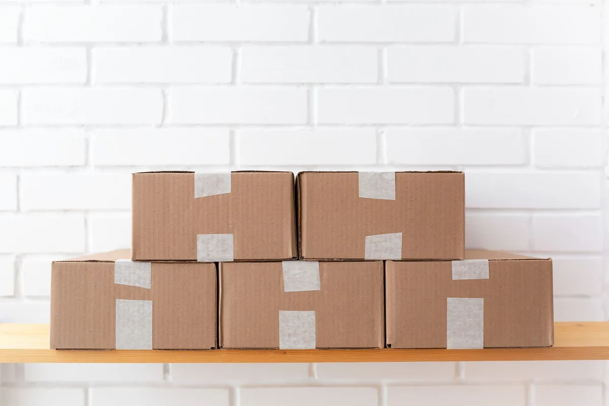 Five taped cardboard parcels stacked on a wooden shelf against a white brick wall