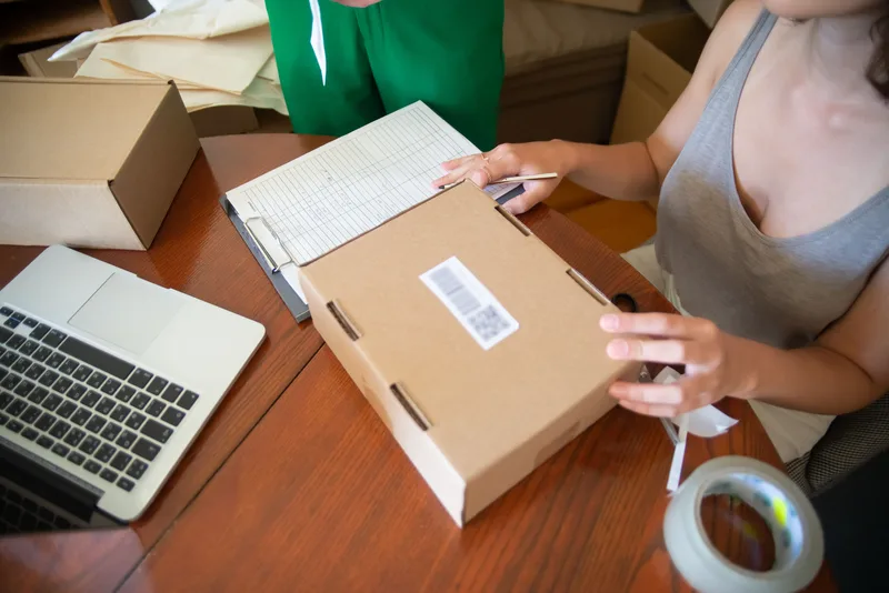 Cardboard box with a QR label on a wooden table beside a laptop, a paper checklist and a roll of tape